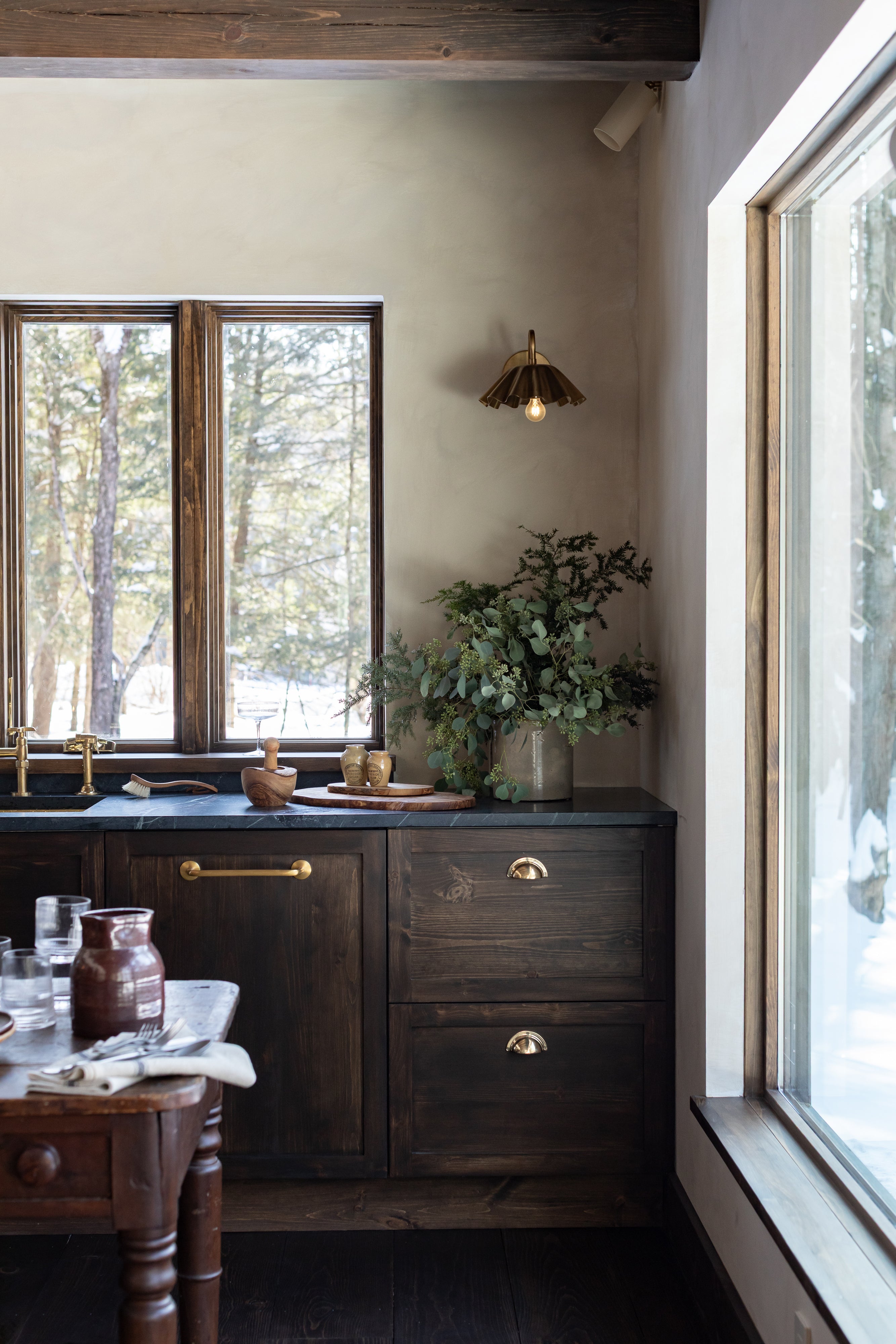 Dark wood kitchen cabinetry with unlaquered brass hardware, soapstone countertop, and windows overlooking a snowy forest.