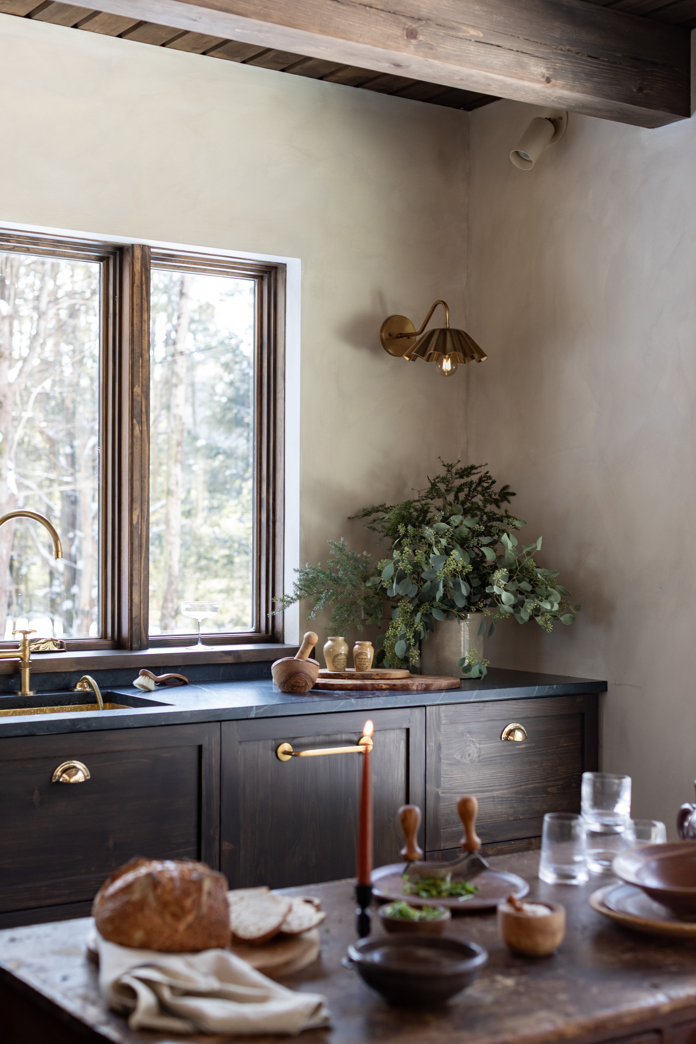 Kitchen featuring dark wood cabinetry, brass hardware, black countertop, and wood-framed windows, with a styled vintage kitchen island in the foreground.